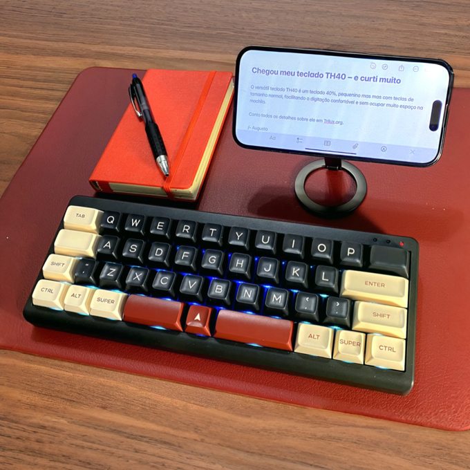 Foto do teclado TH40 na minha bancada, ao lado de um bloco e de uma caneta para permitir percepção da proporção de seu tamanho - a largura é de 25 centímetros.