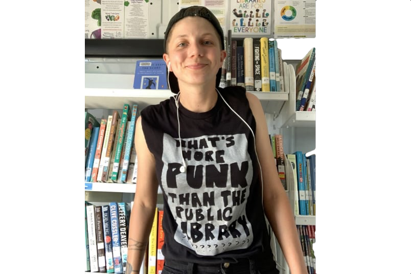 Foto de uma mulher jovem, sorrindo para a câmera, de boné preto virado para trás, com uma camiseta preta onde se lê “What's more punk than the publica library?”, e com tatuagens perto do seu pulso direito.