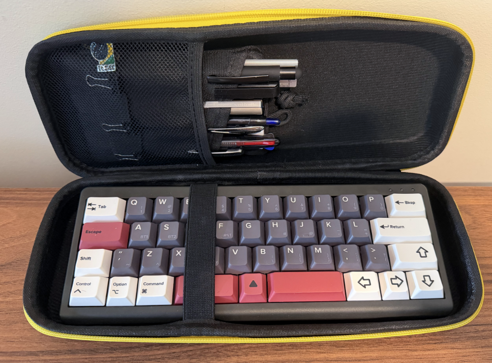 Foto de um teclado em layout 40%, acondicionado em um estojo rígido, com zíper, com um bolso no qual pode ser visto também um estojo de canetas.