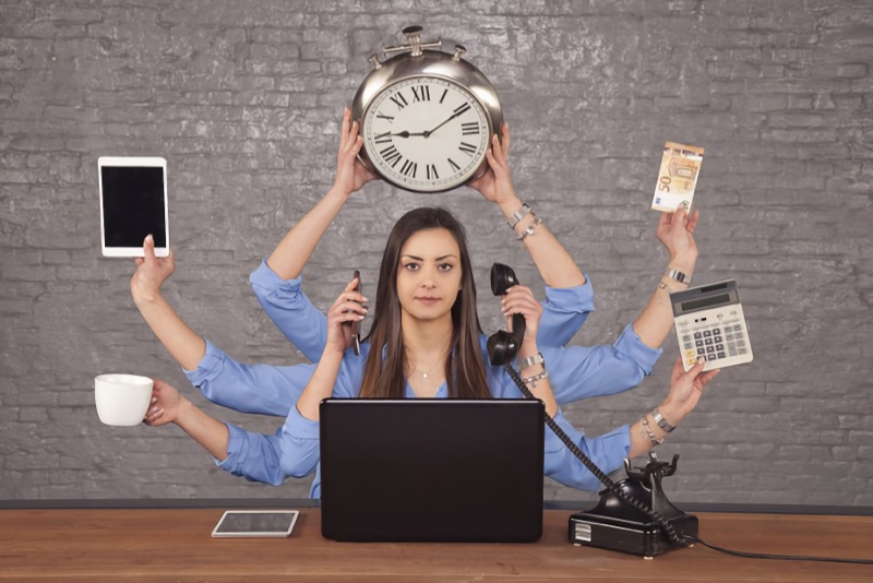 Uma mulher segurando 2 telefones, um relógio de parede, uma calculadora, cédulas, um tablet e uma xícara.