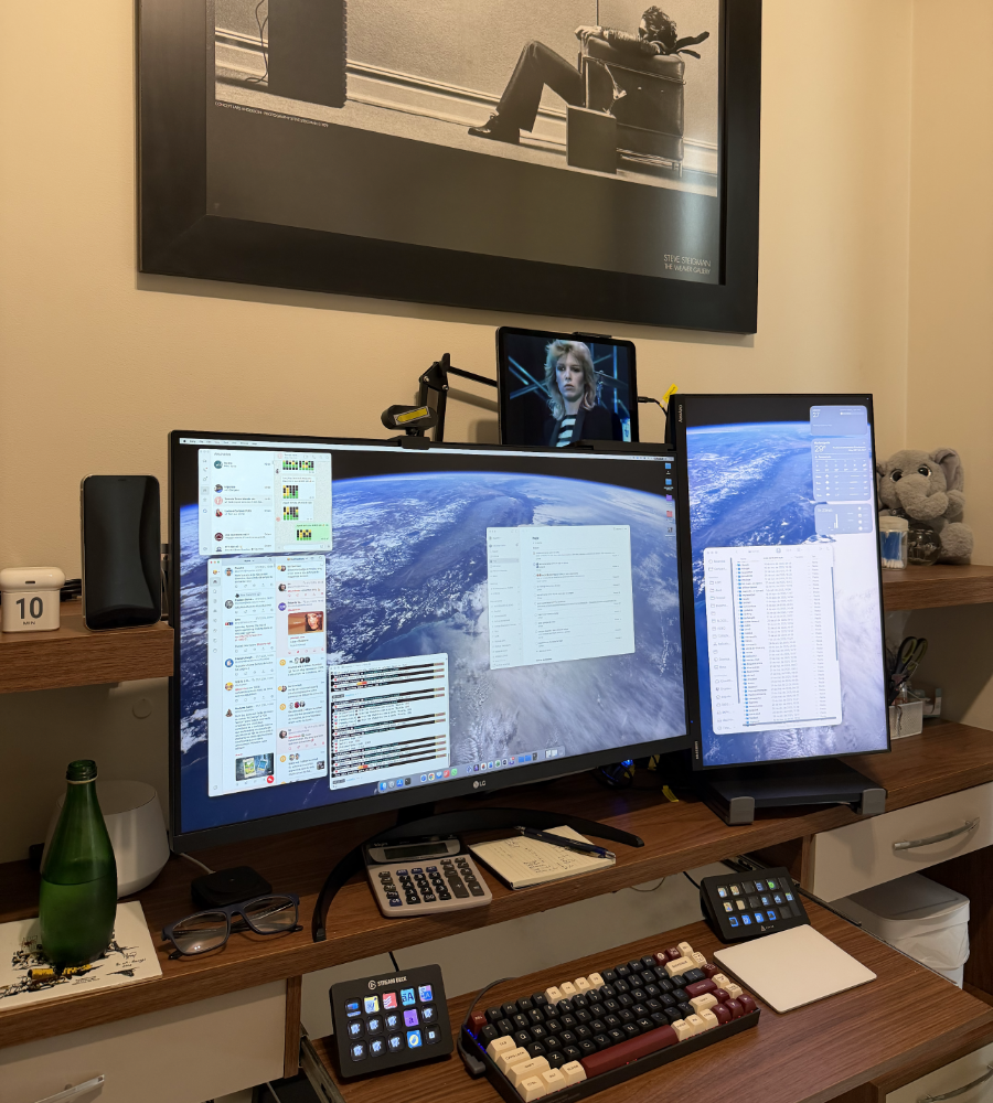 Foto do meu ambiente de trabalho, com 2 monitores lado a lado, uma telinha menor acima de um deles tocando um clip de rock dos anos 80, e um teclado de 84 teclas com teclinhas em uma combinação de cores chamada Rome. Ao lado, uma garrafa de água e um elefante de pelúcia.