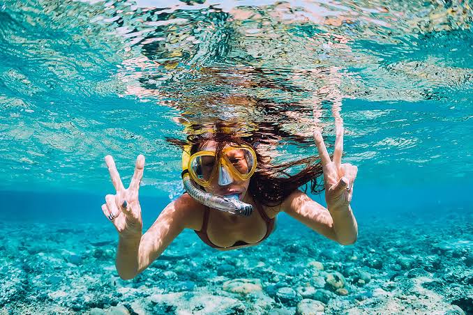 Mulher praticante de snorkel faz sinal de V com os dedos, logo abaixo da superfície da água.