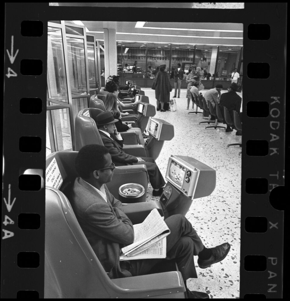 Foto em preto e branco de pessoas assistindo a aparelhos de televisão individuais que funcionam com moedas, no terminal Greyhound Bus em Los Angeles, Califórnia, 1969